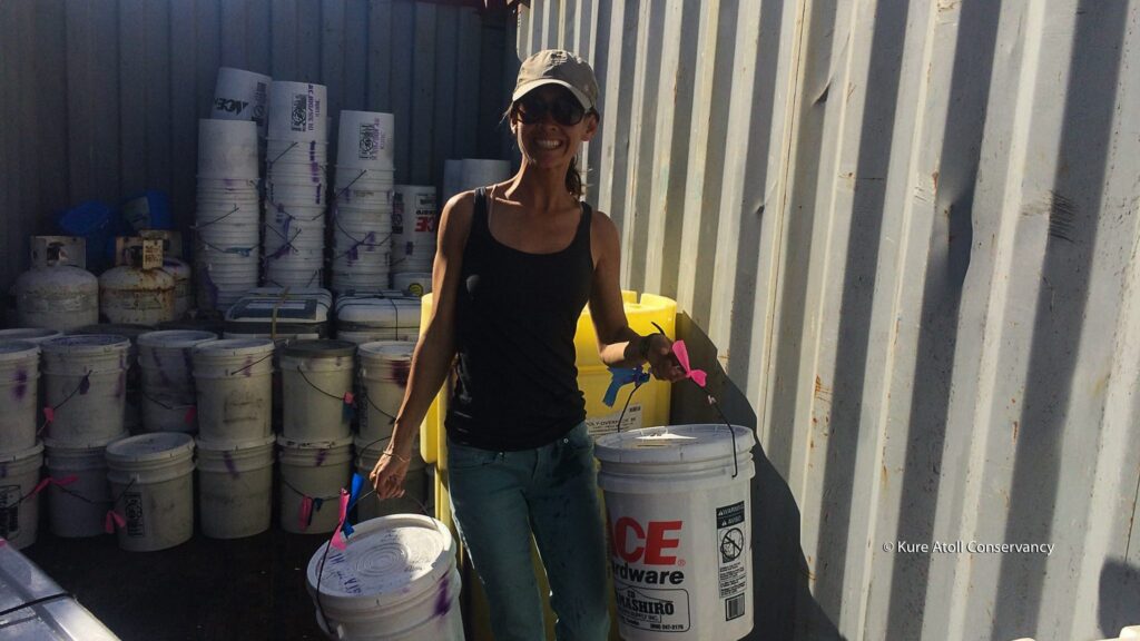 Woman holding buckets and a bag, standing near containers.